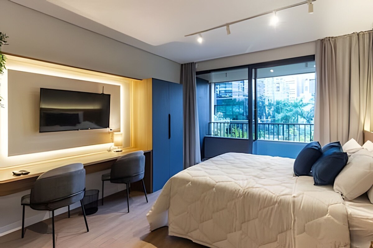 A modern hotel room with a bed, a desk with a chair, a TV, and a large window with a view of buildings and trees.