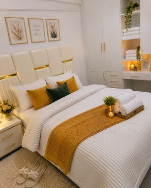 A neatly arranged bedroom with a bed, bedside tables, a nightstand, and a shelf with towels and a small plant.