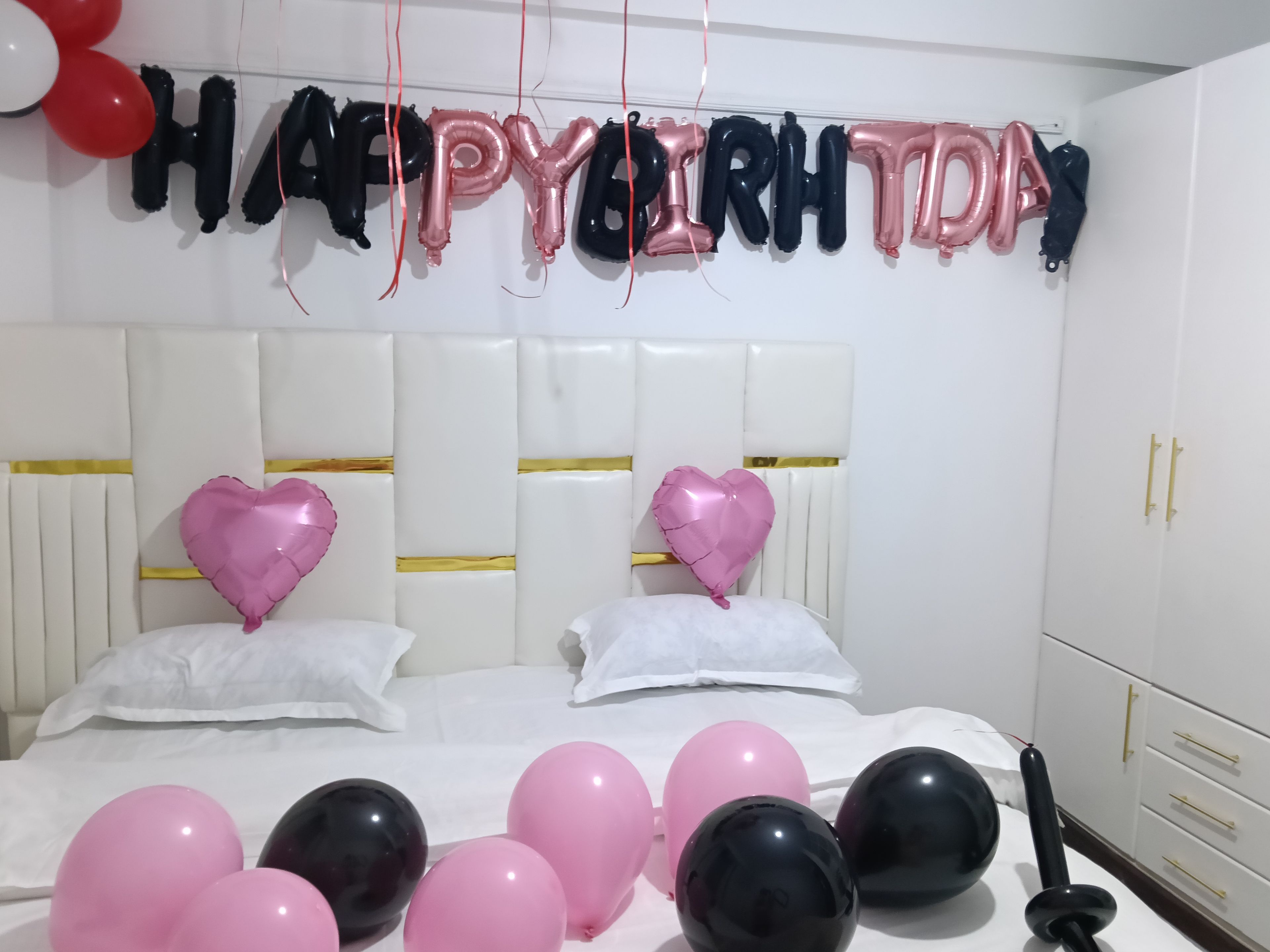 A bedroom with a bed, pillows, and a wall decoration with balloons spelling 'HAPPY BIRTHDAY'.