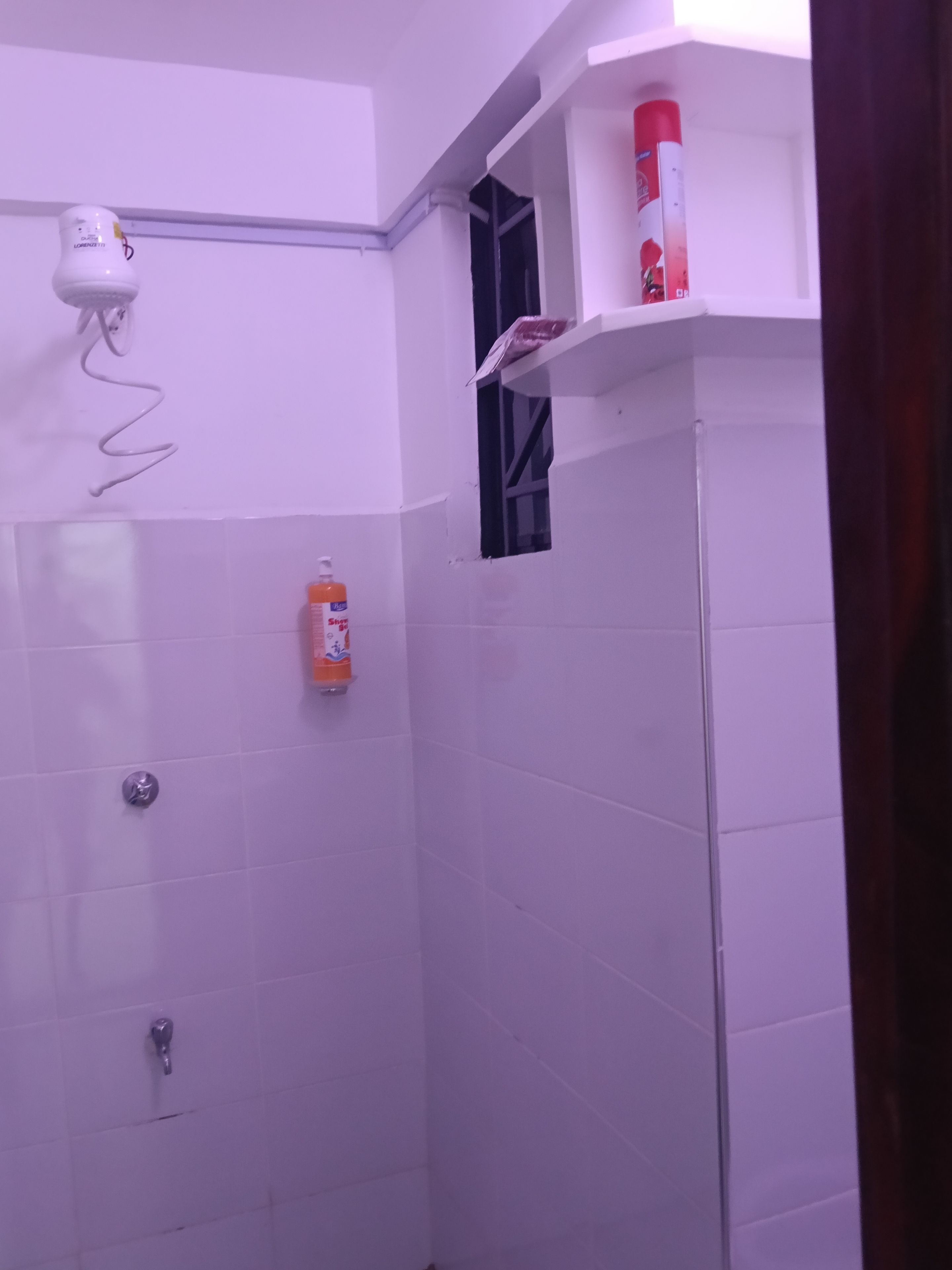 A bathroom with white tiled walls, a shower, a hairdryer, and a shelf with a red bottle.