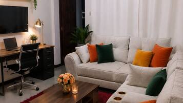 A modern living room with a grey sofa, a wooden coffee table, a red rug, a TV, a desk with a laptop, and a plant.