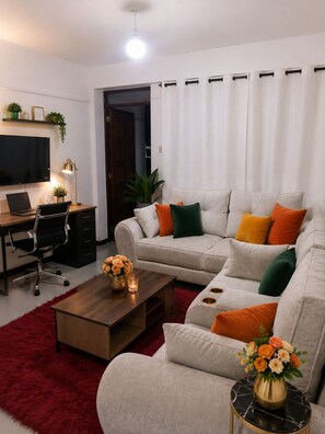 A modern living room with a grey sofa, a wooden coffee table, a red rug, a TV, a desk with a laptop, and a plant. - Cozy 1-bedroom apartment in lovely Nanyuki with AC.Mt Kenya view, Olpejeta Conse (Nanyuki)