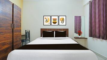 A hotel room with a bed, a wardrobe, a chair, a bedside table with a vase of flowers, and three framed artworks on the wall.