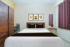 A hotel room with a bed, a wardrobe, a chair, a bedside table with a vase of flowers, and three framed artworks on the wall.