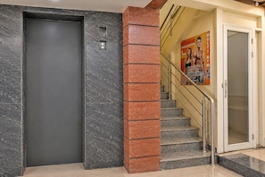 Un ascensor moderno con puerta gris, un pilar de ladrillo rojo y una escalera con puerta de cristal que da a una habitación.