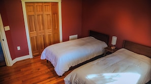 Una habitación con dos camas, suelo de madera, un armario de madera y una mesita de noche con lámpara.