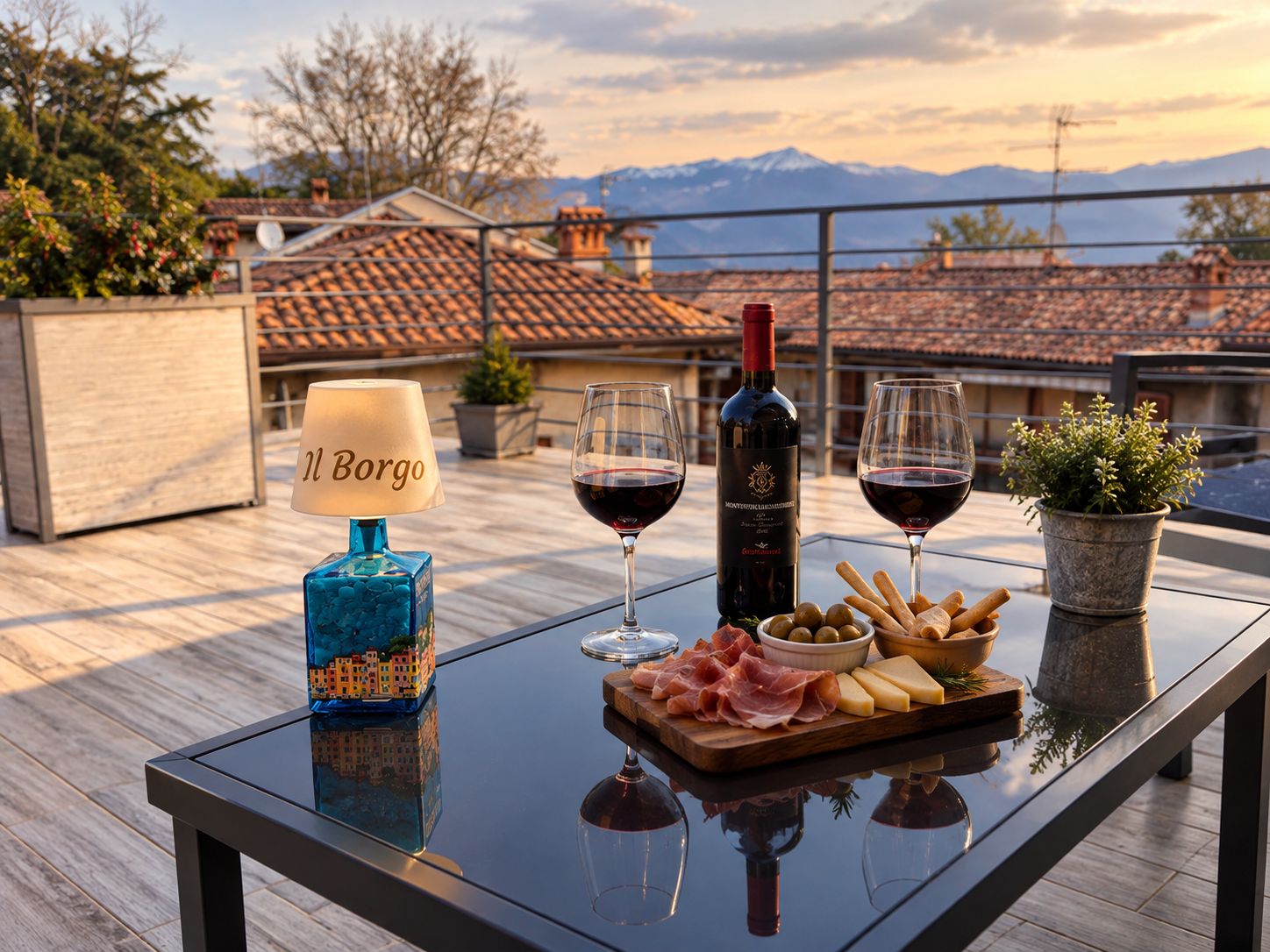 Une table avec une bouteille de vin, des verres, une lampe et une planche à fromages garnie d’olives et de crackers, le tout placé sur un balcon donnant sur une vue panoramique de la ville.