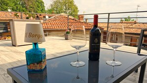Mesa com garrafa de vinho, duas taças de vinho e um abajur em varanda com vista para a paisagem urbana.