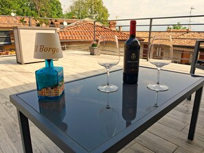 Ein Tisch mit einer Weinflasche, zwei Weingläsern und einer Lampe auf einem Balkon mit Blick auf eine Stadtlandschaft.
