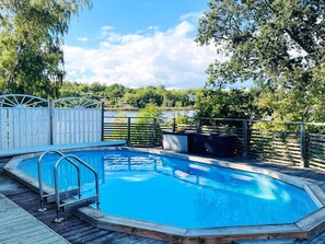 Ein Schwimmbecken mit Holzterrasse, Metallgeländern und Blick auf einen See und Bäume.