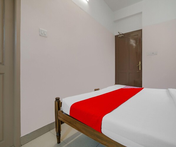 Una cama individual con una colcha roja y blanca en una habitación con paredes blancas y una puerta de madera.