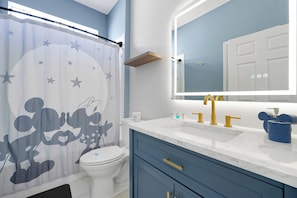 Un baño con cortina de ducha con un personaje de dibujos animados, un inodoro, un lavabo con grifos dorados y un espejo sobre el lavabo.