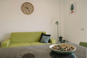 A living room with a green sofa, a decorative wall clock, a framed artwork, a floor lamp, and a table with a bowl of food. - Aetnamare: A 360° View (Mascalucia)