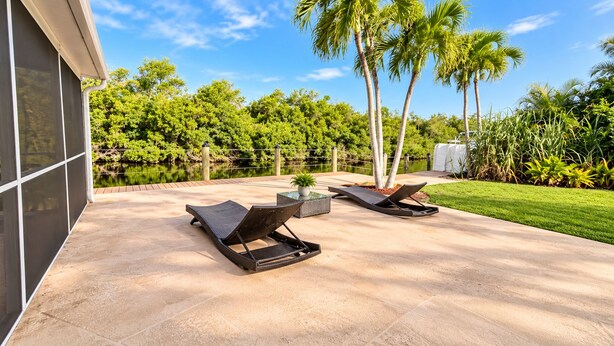 A patio with two lounge chairs and a table, surrounded by a pool and lush greenery. - The Canal Cottage (Fort Myers)