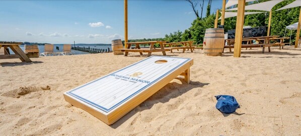 A wooden board game setup on a sandy beach with picnic tables and barrels in the background.