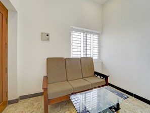 A room with a sofa, a glass coffee table, a window with blinds, and a wall-mounted thermostat. - Premium Royal Care Haven Stays (Sulur)