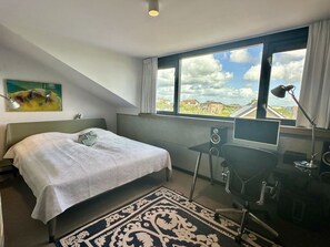 Ein Schlafzimmer mit Bett, Schreibtisch, Computer und Blick auf Gebäude durch ein Fenster.