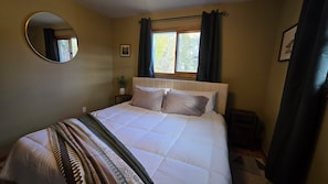 Ein Schlafzimmer mit einem Bett, einem runden Spiegel, einem Fenster mit Vorhängen und einem kleinen Nachttisch mit Pflanze.