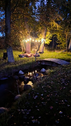 Eine friedliche Außenanlage mit einem kleinen Teich, einer Holzbrücke und Lichterketten, die für eine warme Atmosphäre sorgen.