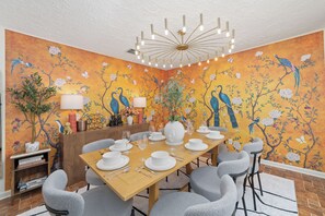 Ein Essbereich mit einem für sechs Personen gedeckten Tisch, umgeben von grau gepolsterten Stühlen. Im Hintergrund ist ein dekoratives Wandbild mit Vögeln und Blumen zu sehen.