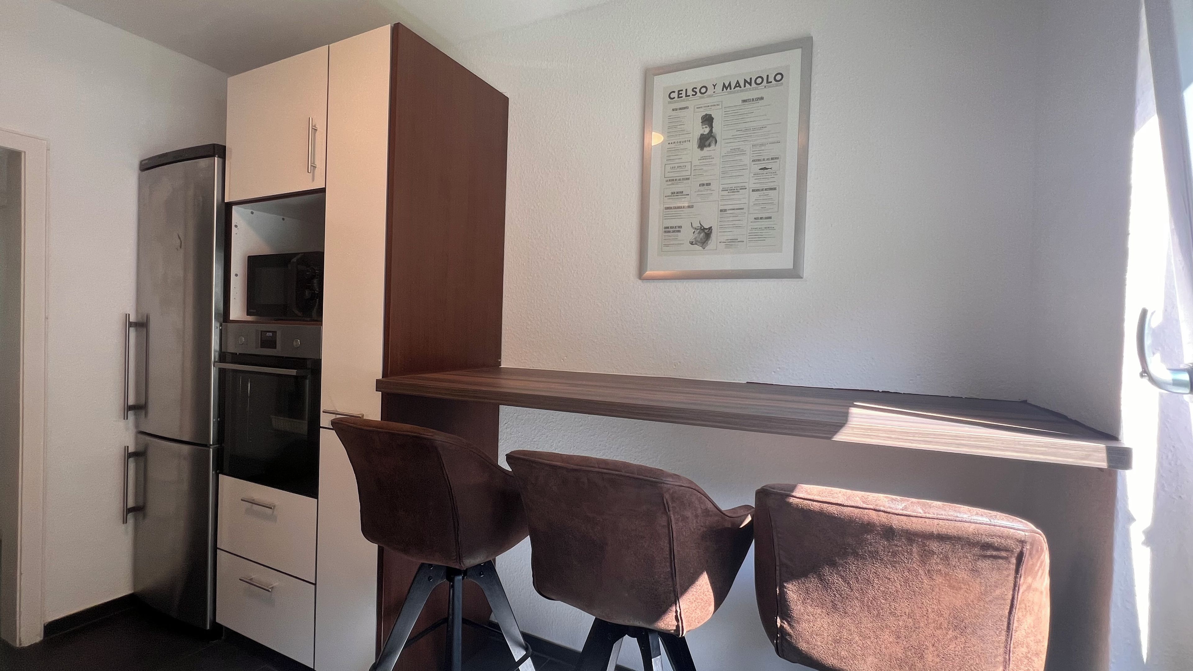 Eine kleine Küchenzeile mit Kühlschrank, Backofen, Mikrowelle und einer Holzarbeitsplatte. Zwei Stühle und eine gerahmte Speisekarte hängen an der Wand.