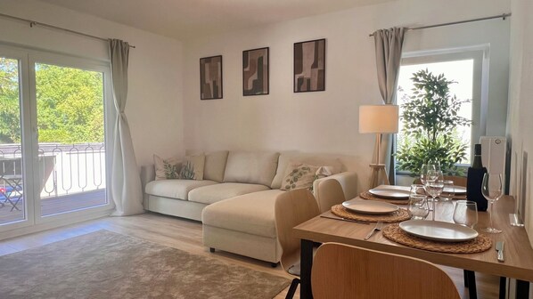 A living room with a sofa, dining table set for four, and a balcony with a view of greenery.