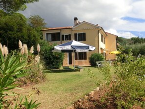 A two-story house with a garden, a patio with a table and chairs, and a large umbrella. - Innamorata Casa Elisio (Capoliveri)