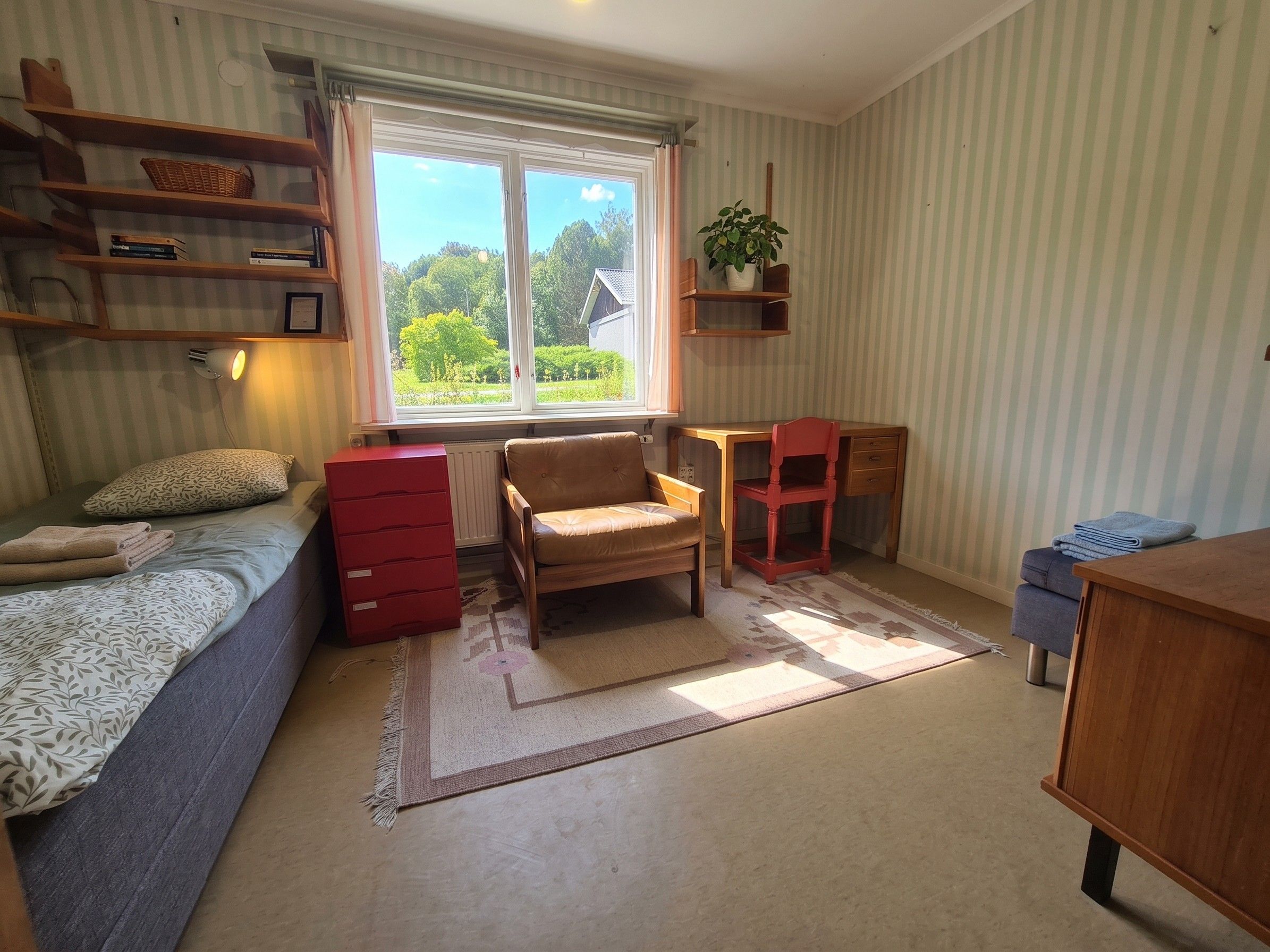 Une pièce comprenant un lit, une commode rouge, une chaise en bois, un bureau et une fenêtre donnant sur un paysage verdoyant.