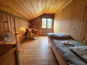A wooden cabin room with a bed, a chair, a fan, and a window.