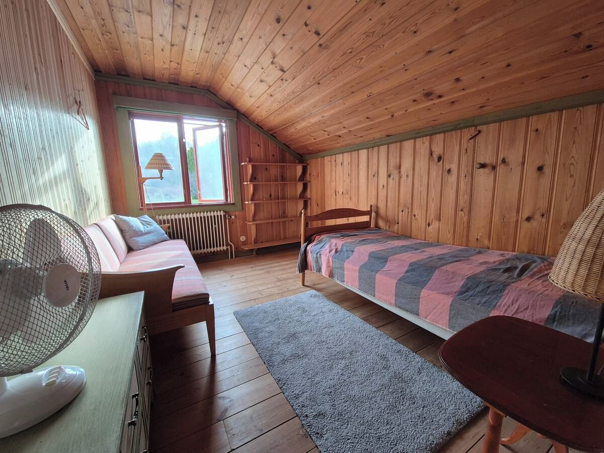Une pièce avec un plafond en pente, des murs en bois, un lit recouvert d’une couverture à rayures, une chaise, une petite table et un ventilateur sur pied.