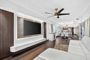 A modern living room with a flat-screen TV, white sofas, a ceiling fan, and a kitchen in the background. - The Baycrest | Heated Pool, Beach and Bay Access (Treasure Island)