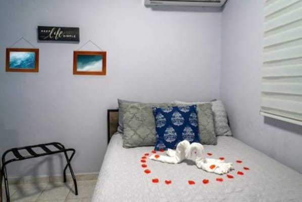 Un dormitorio con cama, almohadas, aire acondicionado de pared y una mesita.