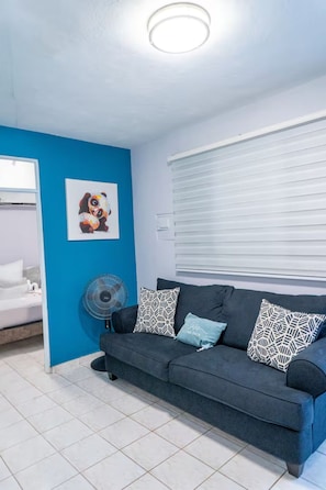 Una habitación con una pared azul de acento, un sofá gris con cojines estampados, un ventilador y una cama con ropa de cama blanca.