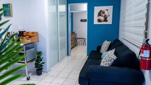 Una habitació amb un sofà negre, una màquina de cafè, una taula petita amb una planta i una porta que dona a un altre espai.
