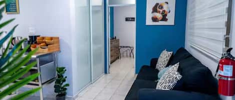 Una habitación con un sofá negro, una cafetera, una mesita con una planta y una puerta que da a otra habitación.