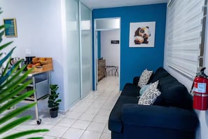 Una habitación con un sofá negro, una cafetera, una mesita con una planta y una puerta que da a otra habitación.