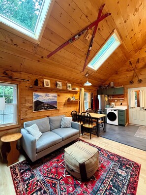 A cozy cabin interior with a wooden ceiling, a sofa, a dining table, and a kitchen area.