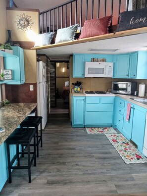 A compact kitchen with teal cabinets, a microwave, and a gas stove. There is a wooden bench and a small island with a sink. - The Cabana (Conway)