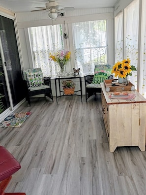 A sunroom with a wooden floor, two chairs with floral cushions, a table with a vase of sunflowers, and a ceiling fan.