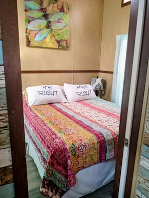 Une chambre à coucher avec un lit, deux oreillers portant les inscriptions « M. Parfait » et « Mme Parfaite », une couvre-lit à motifs et un tableau mural.