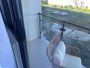 Varanda com cadeira de vime e almofada branca, com vista para um jardim com plantas e um espaço pavimentado.