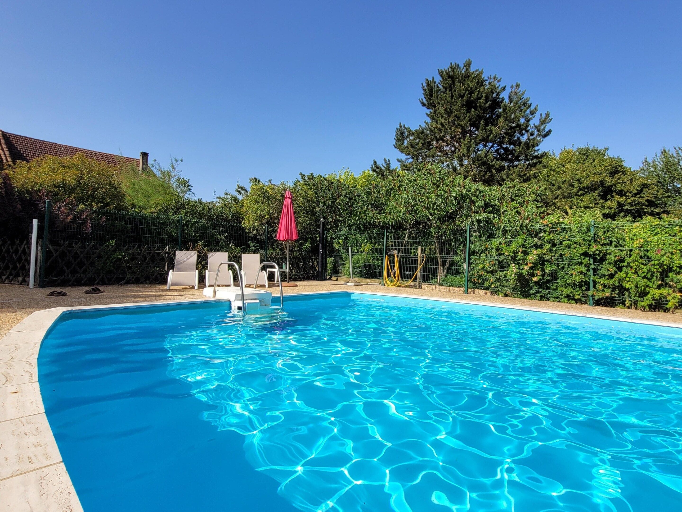 A clear blue swimming pool with a red umbrella, surrounded by a fence and greenery.