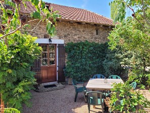 A stone building with a tiled roof, a wooden door with glass panels, and a gravel patio with green chairs. - Authentic Gite With Terrace and Shared Pool (Saint-Saud-Lacoussiere)