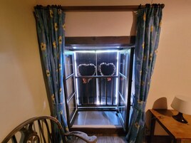Una habitación con ventana de madera, cortinas estampadas con flores, una silla de madera y una mesa con una lámpara.
