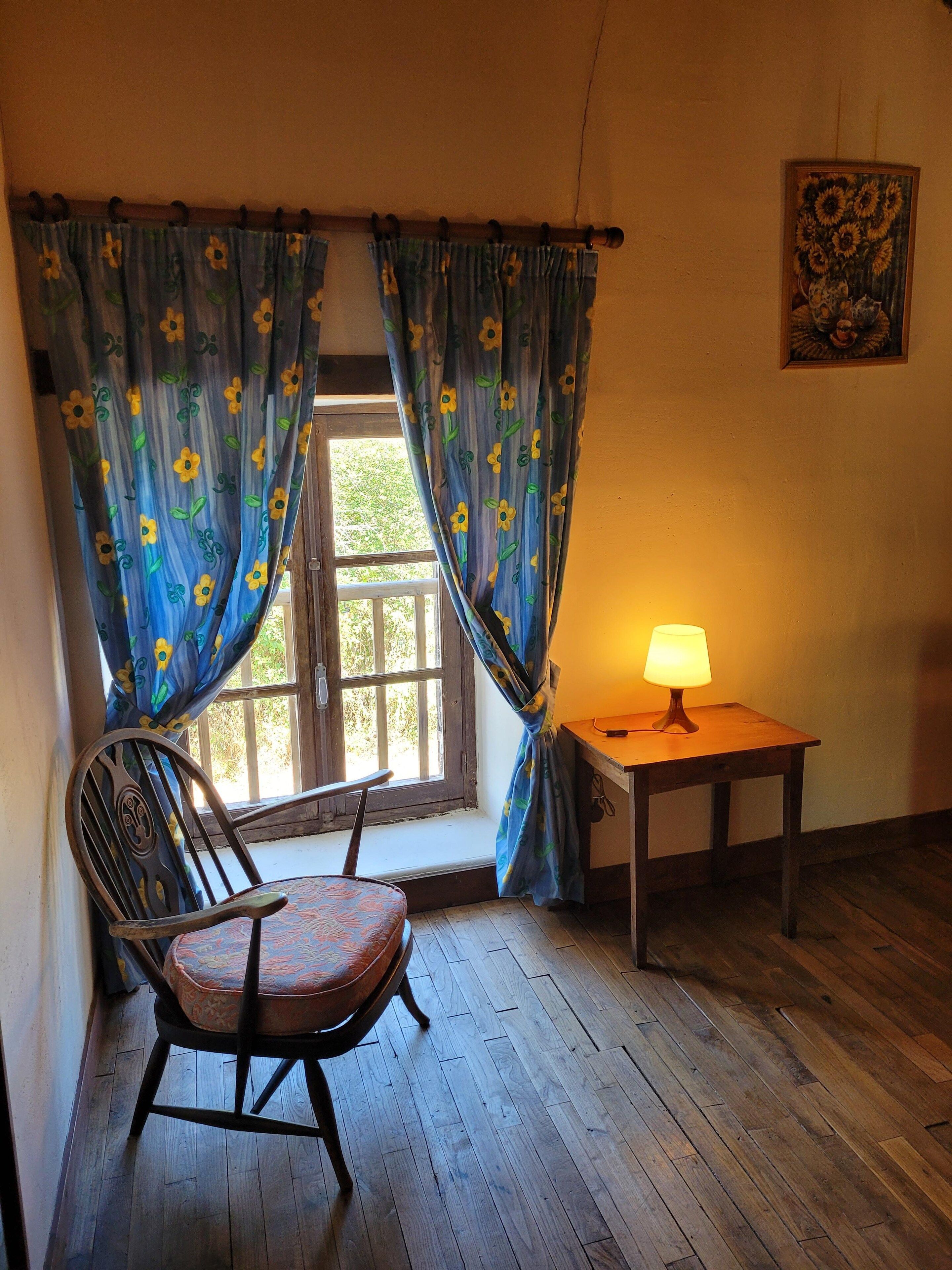 A room with a wooden floor, a chair, a table with a lamp, and a window with curtains.