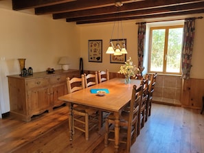 A dining area with a wooden table, chairs, a vase with flowers, and a window with curtains. - Authentic Gite With Terrace and Shared Pool (Saint-Saud-Lacoussiere)