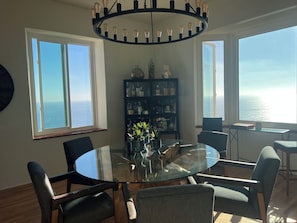 A dining area with a glass table, chairs, a chandelier, a bookshelf, and large windows with a view of the ocean. - Fabulous 3-bedroom house in Laguna Beach with breathtaking panoramic ocean views (Laguna Beach)