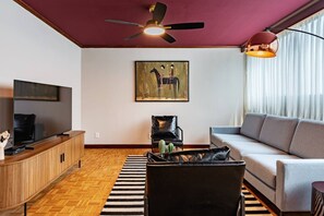Un salon comprenant une télévision, un canapé, un fauteuil, un ventilateur de plafond et un tableau accroché au mur.