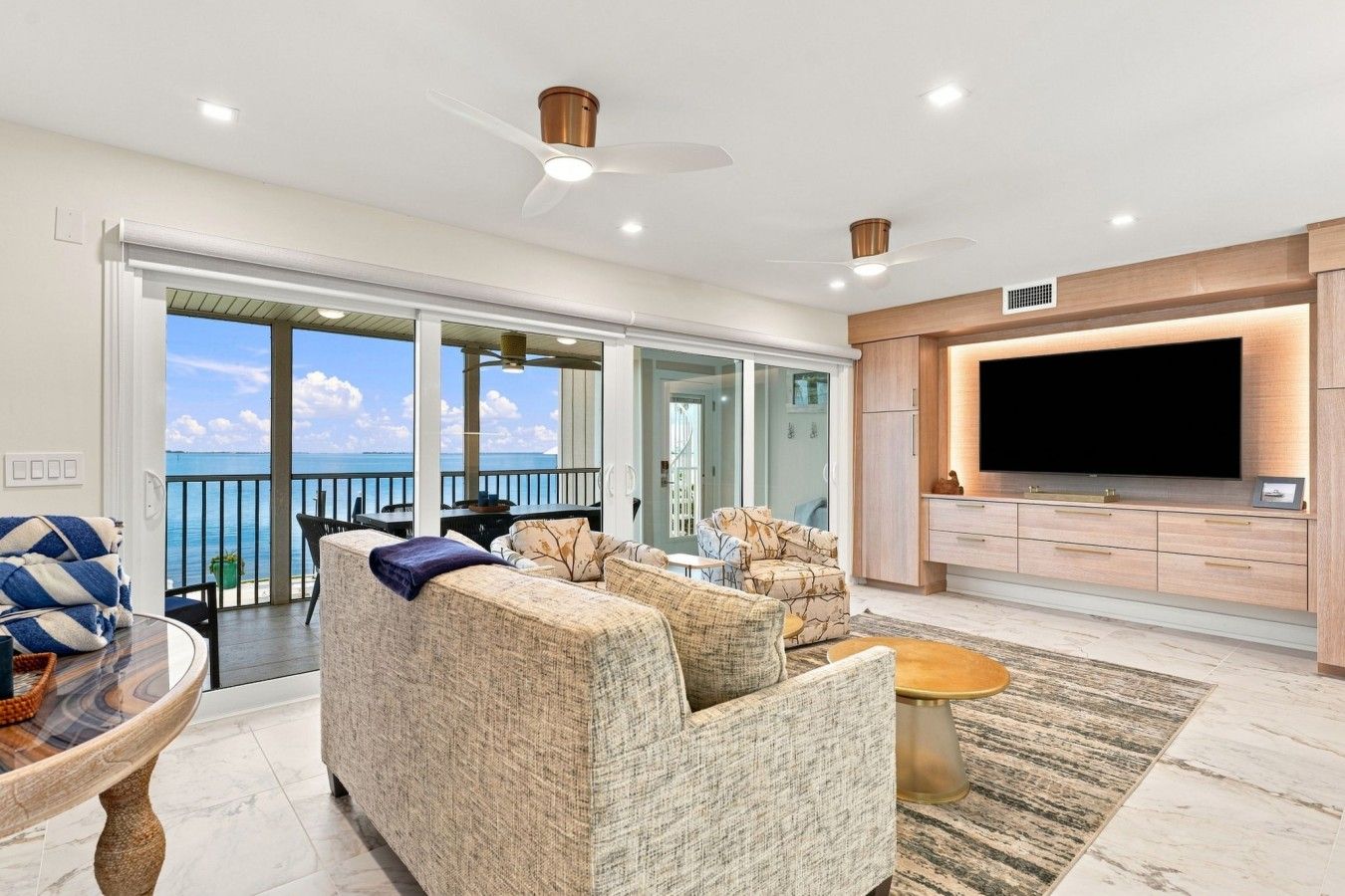 A modern living room with a large TV, comfortable seating, and a view of the sea.