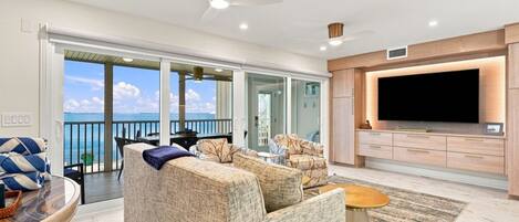 A modern living room with a large TV, comfortable seating, and a view of the sea.
